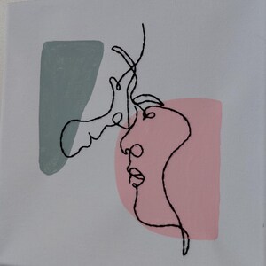 以下が含まれることがあります： キスをする二つの顔のミニマリストな線画が特徴の正方形のキャンバスアート。アートワークは白い背景に黒い糸で描かれ、顔の後ろにはピンクとティールの形があります。キャンバスは壁に取り付けられています。