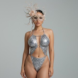 Könnte beinhalten: Ein silberner und grauer Badeanzug mit Schlangenmuster, Neckholder-Top und Cut-Out-Design. Der Badeanzug hat einen hoch geschnittenen Beinausschnitt und ein Ringdetail an der Brust. Das Model trägt eine dekorative Maske und Kopfschmuck mit floralen und Perlenakzenten.