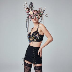 May include: A black crop top with colorful beaded fringe and a matching black skirt with lace-up detailing. The model is wearing a floral and feather mask with ribbons and branches.