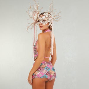 May include: A sequined halter top and matching shorts in pink, purple, and gold geometric patterns. The outfit is paired with an elaborate mask and headpiece adorned with flowers, pearls, and branches. The ensemble is designed for a festive occasion.