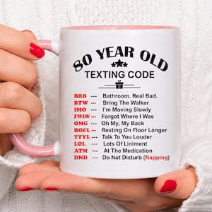 May include: White ceramic mug with a pink interior and handle, featuring the text "80 YEAR OLD TEXTING CODE." The mug lists common texting abbreviations with their meanings, such as "BRB" for "Bathroom. Real Bad." and "DND" for "Do Not Disturb (Napping)."