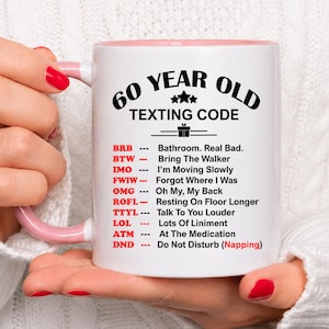 May include: White ceramic mug with a pink interior and handle, featuring the text "60 YEAR OLD TEXTING CODE." The mug lists humorous acronyms like "BRB" and "OMG." Held by a person with red nail polish.