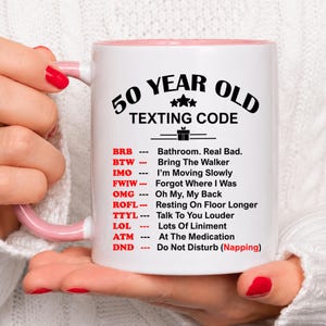 May include: White ceramic mug with a pink interior and handle, featuring the text "50 YEAR OLD TEXTING CODE" and a list of texting acronyms. The mug is held by a person with red nail polish.