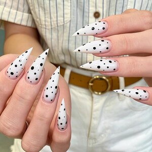 Pode incluir: Unhas em formato stiletto com uma base branca brilhante decorada com pequenos pontos pretos espalhados. As unhas são longas e pontiagudas, exibindo um design monocromático limpo e de alto contraste que cobre toda a superfície.