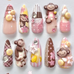 May include: A set of ten almond-shaped press-on nails featuring a variety of designs including brown monkeys, yellow bananas, pink cherry blossoms, white lace patterns, piano keys, musical notes, plaid textures, and small star and circular charms.