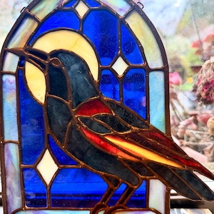 Peut inclure: Un attrape-soleil en vitrail en forme de fenêtre arquée représentant un oiseau noir avec une tache rouge sur l'aile et une plume de queue jaune. L'oiseau est placé sur un fond bleu vif avec des accents en forme d'étoile et une lune jaune pâle.