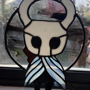 May include: A stained glass suncatcher featuring the character from Hollow Knight. The piece is constructed with white glass for the head and horns, black glass for the eyes and body, and light blue textured glass for the cloak, all held by lead.