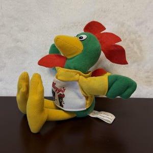 May include: A plush toy of a green rooster with a red comb and wattle, a yellow beak, and yellow feet. The rooster wears a white shirt with the Kellogg's Corn Flakes logo and a graphic of a rooster head. A white fabric tag is attached to the side.