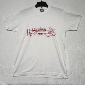 May include: A white short-sleeved t-shirt featuring a red graphic print on the chest. The design includes a musical staff with a treble clef, the text Gingham Cloggers, and an illustration of a pair of shoes. The shirt has a crew neckline.