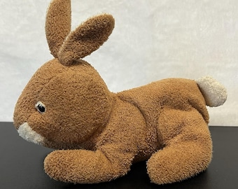 May include: A brown plush rabbit toy with a textured, fuzzy fabric finish. The stuffed animal features long upright ears, a small white nose patch, a white tail, and dark eyes, depicted in a resting, lying-down position with a soft, rounded body.