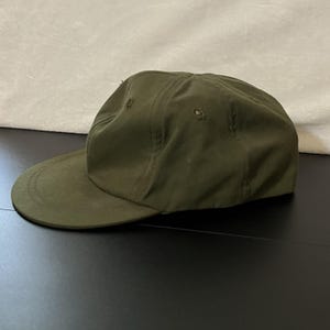 May include: An olive green baseball cap featuring a structured crown with stitched eyelets and a flat, rounded brim. The cap is constructed from a solid-colored fabric with visible seams running along the panels, creating a clean and uniform appearance.