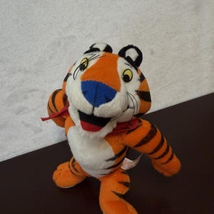 May include: A plush tiger toy featuring orange fur with black stripes, a white chest and face, a blue nose, and a red bandana around its neck. The character has yellow eyes, black ears, and a wide, smiling mouth with a visible red tongue.