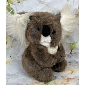 May include: A plush koala toy featuring dark brown fur, large fluffy white ears, and a prominent black nose. The stuffed animal has a white patch on its chest and face, with dark eyes positioned above the nose, crafted with a soft texture.