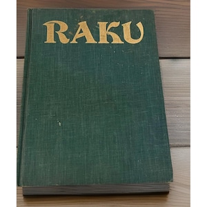 May include: A hardcover book with a dark green, textured fabric cover. The word RAKU is printed in large, gold, stylized capital letters across the upper portion of the front cover. The book has a visible spine and thick, white paper pages.