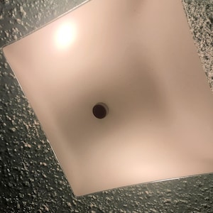 Puede incluir: Una lámpara de techo cuadrada empotrada con un panel de vidrio esmerilado. La luz emite un brillo suave, y un elemento circular marrón oscuro está centrado en el panel. La lámpara está colocada contra una pared texturizada de color gris oscuro.