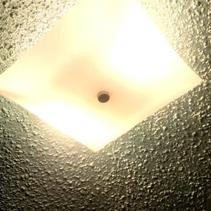 Puede incluir: Un plafón cuadrado empotrado con un panel de vidrio esmerilado. La luz emite un brillo cálido y suave. El techo circundante tiene un acabado texturizado de color gris oscuro.