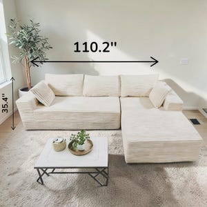 May include: Cream-colored sectional sofa with corduroy texture. The sofa is 110.2 inches long and 35.4 inches high. Includes matching throw pillows. A white square coffee table with a decorative top and a small plant is in front of the sofa.