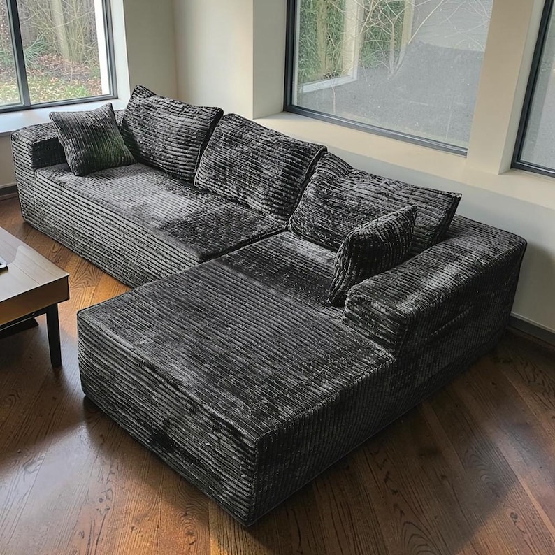 May include: A large, sectional sofa with a dark gray corduroy fabric. The sofa has multiple cushions and a chaise lounge on one end. The room has hardwood floors and large windows.