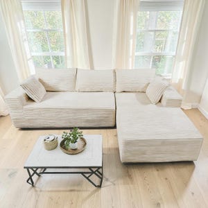 May include: Cream-colored sectional sofa with corduroy texture, featuring multiple cushions and an attached chaise lounge. A white square coffee table with a black metal base and a small plant on top completes the living room setting.
