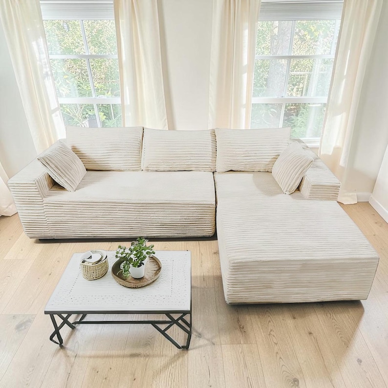 May include: Cream-colored sectional sofa with corduroy texture, featuring multiple cushions and an attached chaise lounge. A white square coffee table with a black metal base and a small plant on top completes the living room setting.