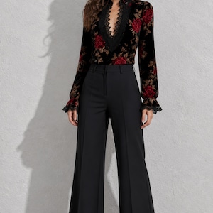 Puede incluir: Una blusa de terciopelo negro de manga larga con un escote en V profundo con ribete de encaje negro y un estampado floral de rosas rojas, combinada con pantalones negros de talle alto y pernera ancha con una raya marcada en el centro.
