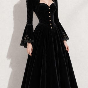 May include: A floor-length black velvet dress featuring long bell sleeves with lace trim and gold floral embroidery. The bodice includes a sweetheart neckline, a row of small gold buttons down the front, and structured shoulders with matching embroidery.