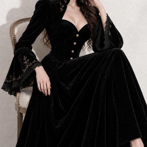 May include: A floor-length black velvet dress featuring a sweetheart neckline, a high collar, and long flared sleeves with intricate gold embroidery and black lace trim. The bodice is fitted with a row of small gold buttons down the center front.