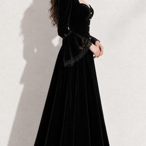 May include: A floor-length black velvet gown featuring a high collar, long sleeves with flared cuffs, and a row of buttons down the front bodice. The garment is accented with delicate gold embroidery along the collar and cuffs, and black lace trim.