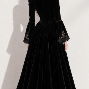 May include: A floor-length black velvet gown featuring a high collar, long sleeves with flared cuffs, and intricate gold embroidery detailing on the lower sleeves. The dress has a fitted bodice and a full, flowing skirt that reaches the floor.