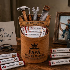 May include: A wooden Father's Day gift set with a toolbox-shaped container holding tools and chocolate bars. A framed photo, a card that says "Happy Father's Day!", and a pair of glasses are also on the table. The container has the word "Papa" on it.