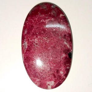 May include: An oval-shaped cabochon gemstone featuring a deep magenta and crimson color palette with mottled white and grey inclusions throughout the surface. The stone has a smooth, polished finish with a single small black speck on the right side.