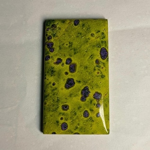 Könnte beinhalten: Ein rechteckiger Cabochon mit einer glatten, polierten Oberfläche. Die Grundfarbe ist ein leuchtendes Limettengrün mit einer durchgehend gesprenkelten Textur. Über die Oberfläche sind unregelmäßige, dunkelviolette Flecken verteilt.