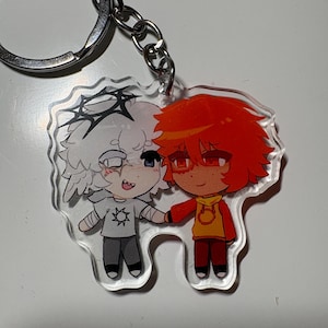 Puede incluir: Un llavero de acrílico transparente con dos personajes de dibujos animados tomados de la mano. Un personaje tiene el pelo blanco con una corona negra y una camiseta gris con un icono de sol. El otro tiene el pelo naranja brillante y una sudadera amarilla y naranja.