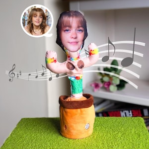 May include: A novelty plush cactus toy with a human face, wearing a floral lei and a brown pot. The toy has arms with colourful accents and musical notes floating around it. A photo of a woman is in a speech bubble.
