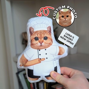 May include: A custom-shaped plush pillow featuring a ginger cat wearing a white chef hat and uniform, holding a wooden rolling pin. A small white sign attached to the side reads LUNA'S BAKERY NOW OPEN! A circular inset shows the original cat photo.