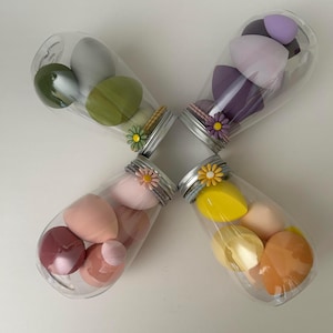 May include: Four clear plastic jars filled with colourful makeup sponges. Each jar is decorated with a daisy and a decorative band around the lid. The sponges are in shades of green, purple, pink, and yellow.