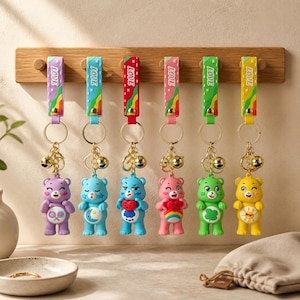 May include: A selection of bear-shaped keyrings in various colours, suspended from a wooden rack. Each keyring showcases a different coloured bear with unique designs and a gold bell charm. The straps are printed with the word "LOVE".