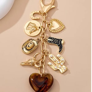 May include: Gold-toned keychain with multiple charms. Charms include a horseshoe, heart, cowboy hat, cactus, and a heart-shaped pendant. The cactus charm has faux pearl accents. The keychain is attached to a gold clasp.