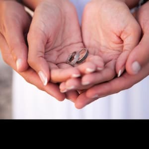 Può includere: Due fedi nuziali, una con pietre chiare e l'altra con pietre scure, sono tenute in mani a coppa. Un'altra coppia di mani le culla, simboleggiando amore e impegno. Gli anelli sono il punto focale dell'immagine.