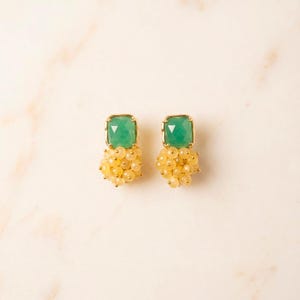 Puede incluir: Un par de pendientes de botón con una piedra preciosa verde de corte cuadrado engastada en metal dorado, con un racimo de pequeñas cuentas redondas de color amarillo pálido suspendidas bajo cada piedra, creando un aspecto texturizado y multicapa.