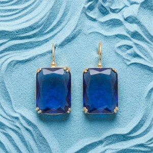 Puede incluir: Un par de pendientes dorados con grandes piedras preciosas rectangulares de color azul oscuro. Los pendientes tienen un diseño de gancho simple y están colocados sobre un fondo azul claro texturizado.