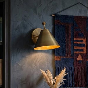May include: A brass wall sconce with a conical shade is mounted on a dark blue wall. The sconce illuminates the wall and a small table with a vase of dried flowers and a stack of books. A woven wall hanging is also visible.