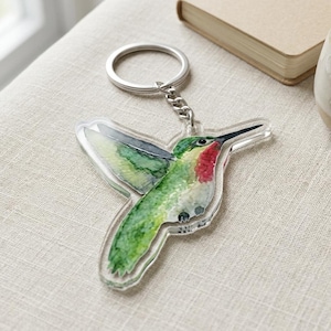 May include: A clear acrylic hummingbird keychain with a silver keyring and chain. The hummingbird is painted with watercolor in shades of green, red, and black. The keychain is resting on a beige linen surface.