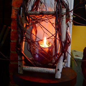 May include: A candle burning inside a rustic twig lantern. The lantern is made of natural branches and has a whitewashed finish. The candle is surrounded by a warm glow.