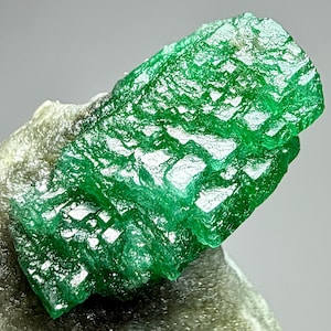 Pode incluir: Uma aproximação de um vibrante grupo de cristais de esmeralda verde. A esmeralda tem uma estrutura cristalina com uma superfície áspera e texturizada. A gema é colocada sobre um fundo neutro e claro, destacando sua cor vívida e forma natural.