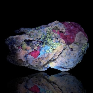 May include: A mineral specimen featuring a rough, irregular texture with vibrant patches of fluorescent pink, bright yellow-green, and muted blue-gray tones across its surface, set against a solid black background with a reflection below.
