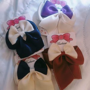 Può includere: Una collezione di fiocchi per capelli in raso in vari colori, tra cui blu navy, bordeaux e viola, ciascuno con un fiocco avorio a contrasto. I fiocchi sono attaccati a piccole carte con la scritta "Fashion".