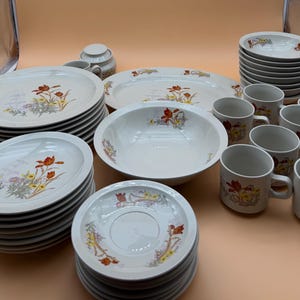 May include: A large dinnerware set featuring white ceramic plates, bowls, mugs, and saucers decorated with a floral pattern of orange, yellow, and green wildflowers. The collection includes stacked dinner plates, salad plates, a serving platter, a bowl, and multiple handled mugs.