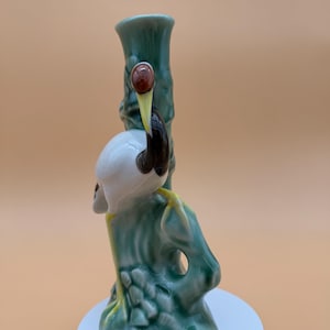 Puede incluir: Figura de cerámica de un pájaro posado en un tronco de árbol verde. El pájaro tiene la cabeza blanca, pico negro y ojos marrones. El tronco del árbol es de color verde claro con detalles amarillos. La figura está sobre un pedestal blanco.