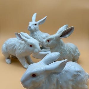 Op de afbeelding: Vier witte keramische konijnenbeeldjes met rode ogen. De konijnen staan in verschillende poses, sommige zitten en andere staan. De beeldjes hebben gedetailleerde vachttexturen en staan tegen een lichtoranje achtergrond.